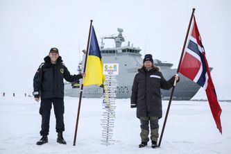 Two people holding flags, standing on snow in front of a ship with a signpost indicating the North Pole.