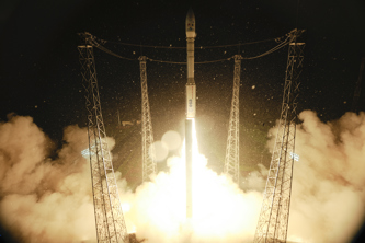 Rocket launch at night, with flames and smoke illuminating the area, surrounded by four tall support towers.