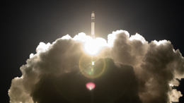 Rocket ascending into the sky, surrounded by bright flames and thick clouds of smoke, against a dark background.